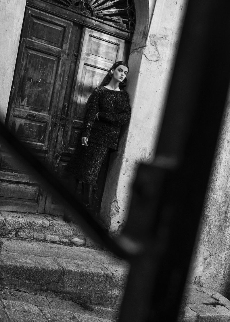 Monochrome fashion editorial image of a woman leaning against an old wooden door in a stone entryway.