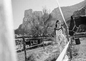 Black and white editorial photograph of a model in a patterned dress standing near a horse by a paddock with mountains in the background