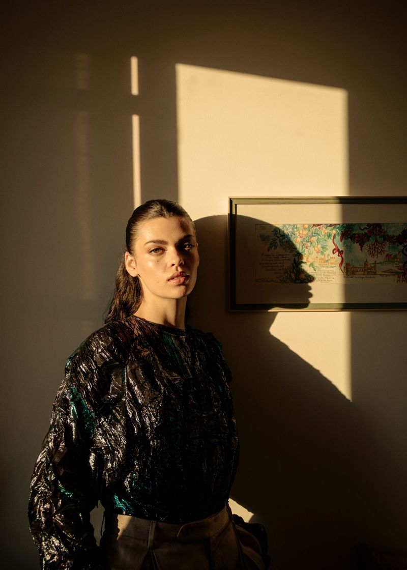 Model with dark hair wearing a textured dark top posing in warm sunlight against an interior wall with a framed picture