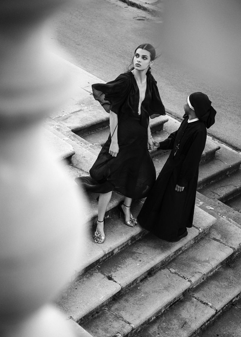 Monochrome fashion editorial image of a woman in a dark dress standing on stone stairs with a companion in religious habit.