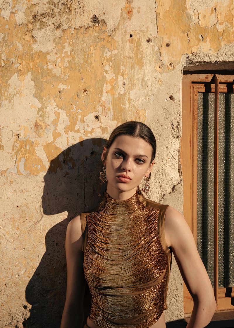 Model wearing a gold textured top standing against a weathered stone wall with a grated window in warm sunlight.