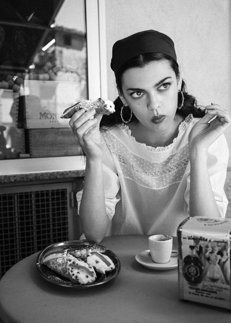 Black and white portrait of a woman in a cafe holding a cannoli at a round table with a small espresso cup.