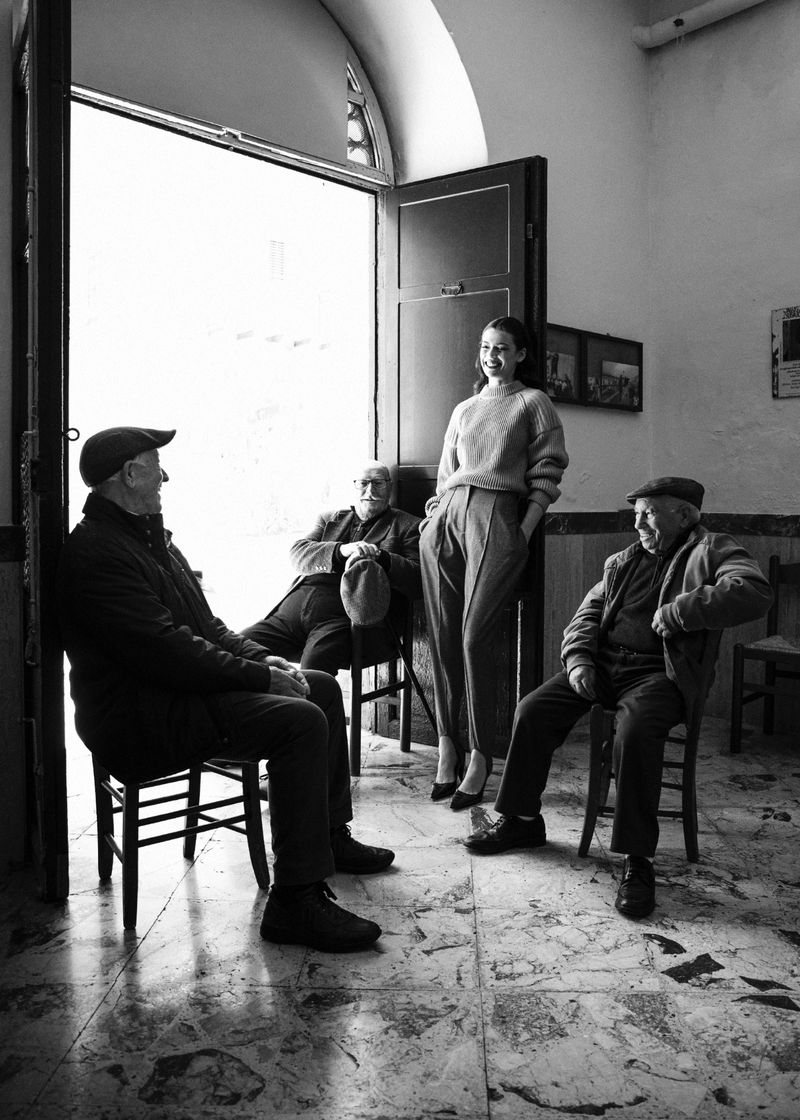 Black and white editorial portrait of a woman standing in a doorway flanked by three seated elderly men in caps
