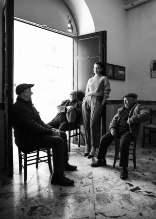 Black and white editorial portrait of a woman standing in a doorway flanked by three seated elderly men in caps