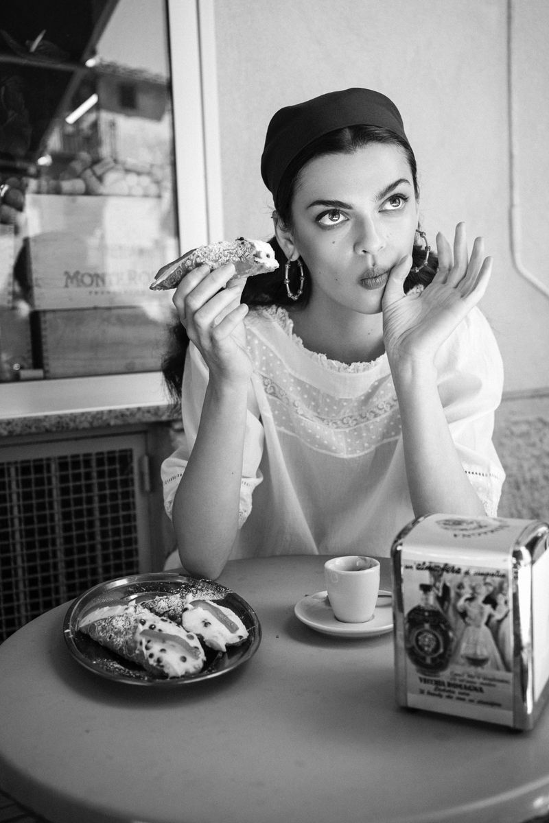 Model seated at an outdoor café table holding a pastry in a black and white fashion editorial by Edwin S Freyer for 5ELEVEN Magazine.