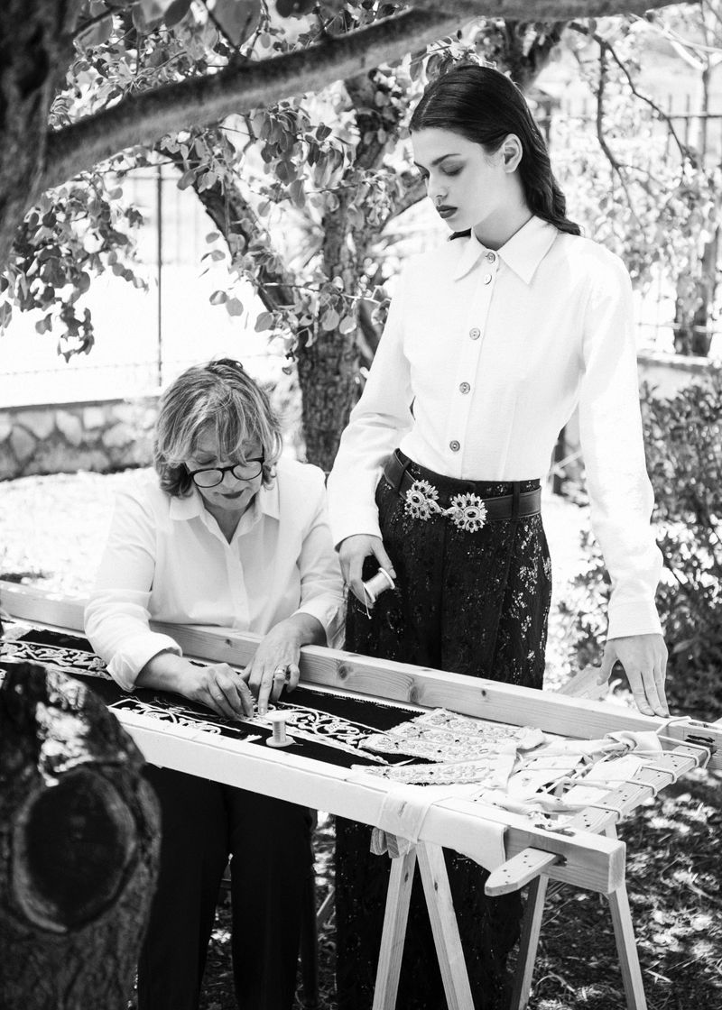 Black and white fashion editorial showing an older woman working at an embroidery frame next to a standing young female model outdoors.