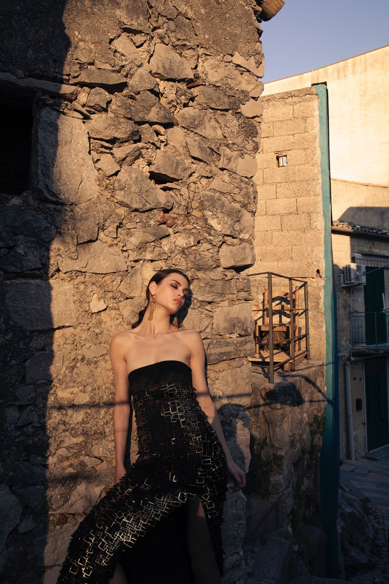 Model leaning against a weathered stone wall wearing a strapless black dress under warm evening sunlight.