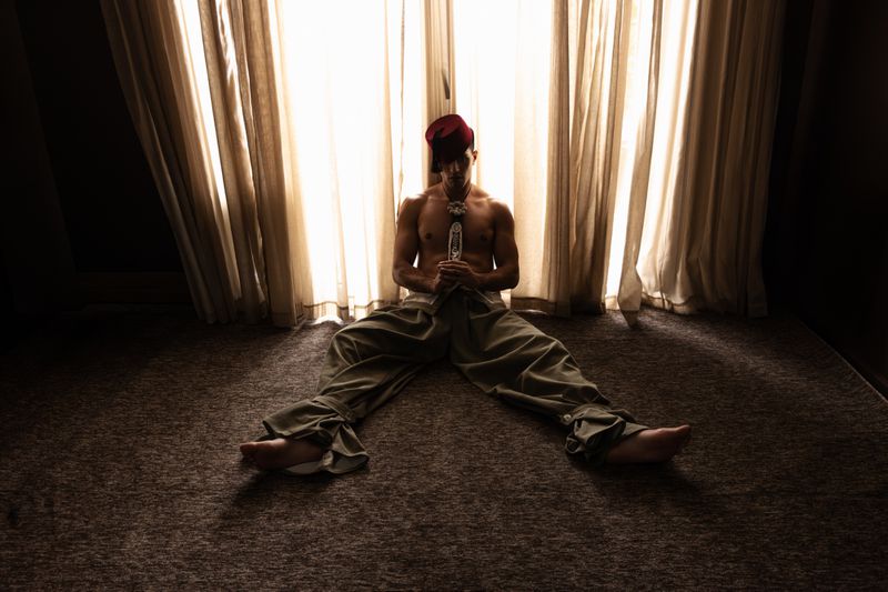 Shirtless male model wearing a red fez and loose trousers sitting on a carpeted floor in front of sheer lit curtains.