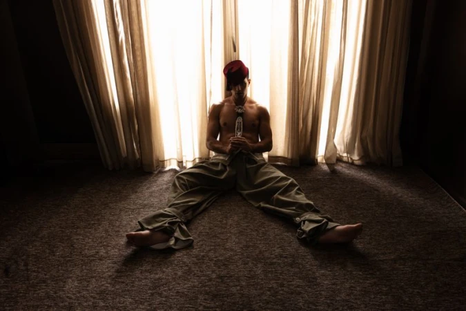 Shirtless male model wearing a red fez and loose trousers sitting on a carpeted floor in front of sheer lit curtains.