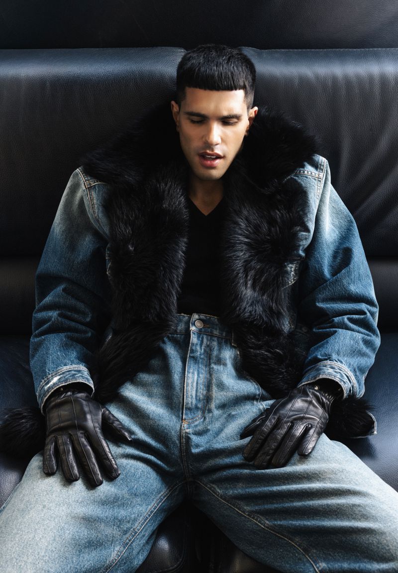 Man wearing a denim jacket with faux fur trim and blue jeans seated on a dark leather couch