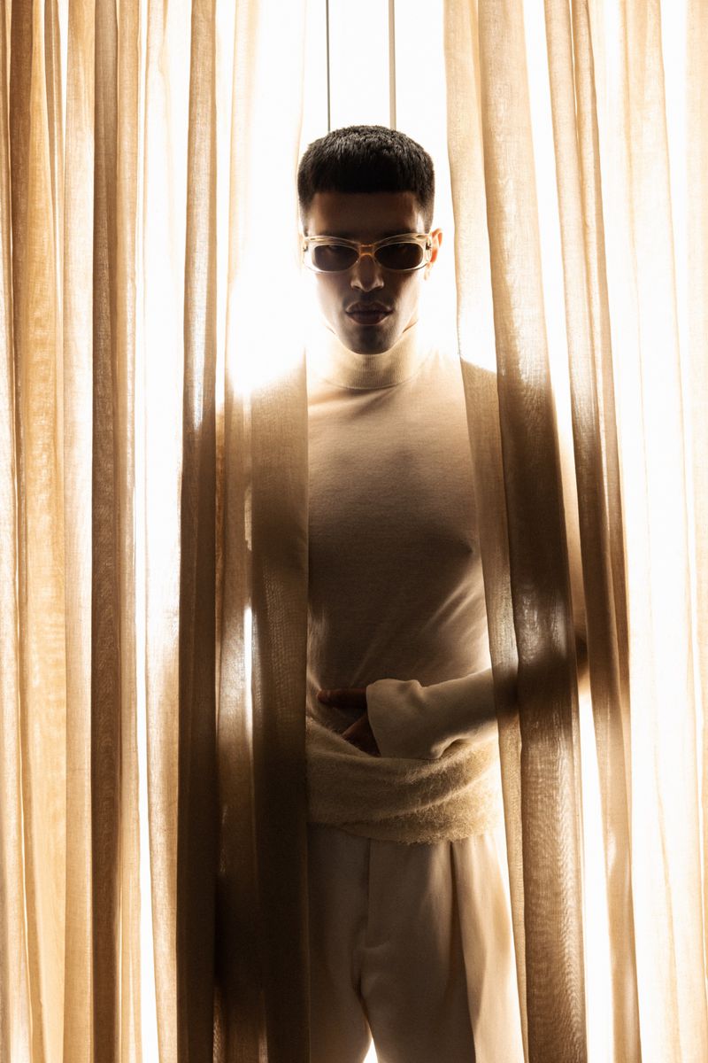 Model Denis Jovanovic wearing sunglasses and light knitwear standing behind sheer vertical window curtains.