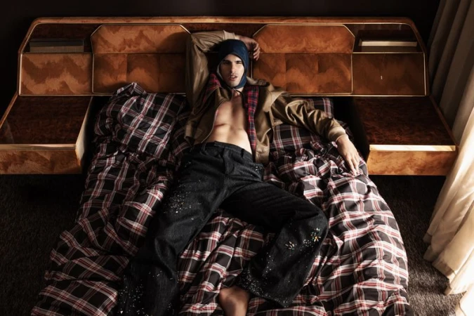 Male model lying on a plaid-covered bed wearing dark trousers, an open brown jacket, and a blue hood.