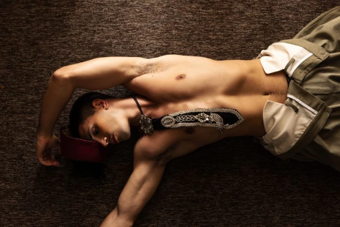 Shirtless male model lying on textured carpet wearing high waisted trousers, embellished neckpiece and fez