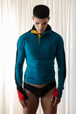 Model wearing a long-sleeved teal, black, and red pullover with dark briefs standing indoors against white curtains
