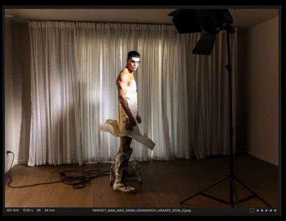 Model Denis Jovanovic wearing a white paper garment and tall boots in a photo studio with white curtains for The Perfect Man Magazine.