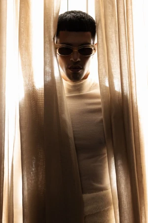 Man wearing sunglasses and a turtleneck sweater peering between sheer curtains for The Perfect Man Magazine.