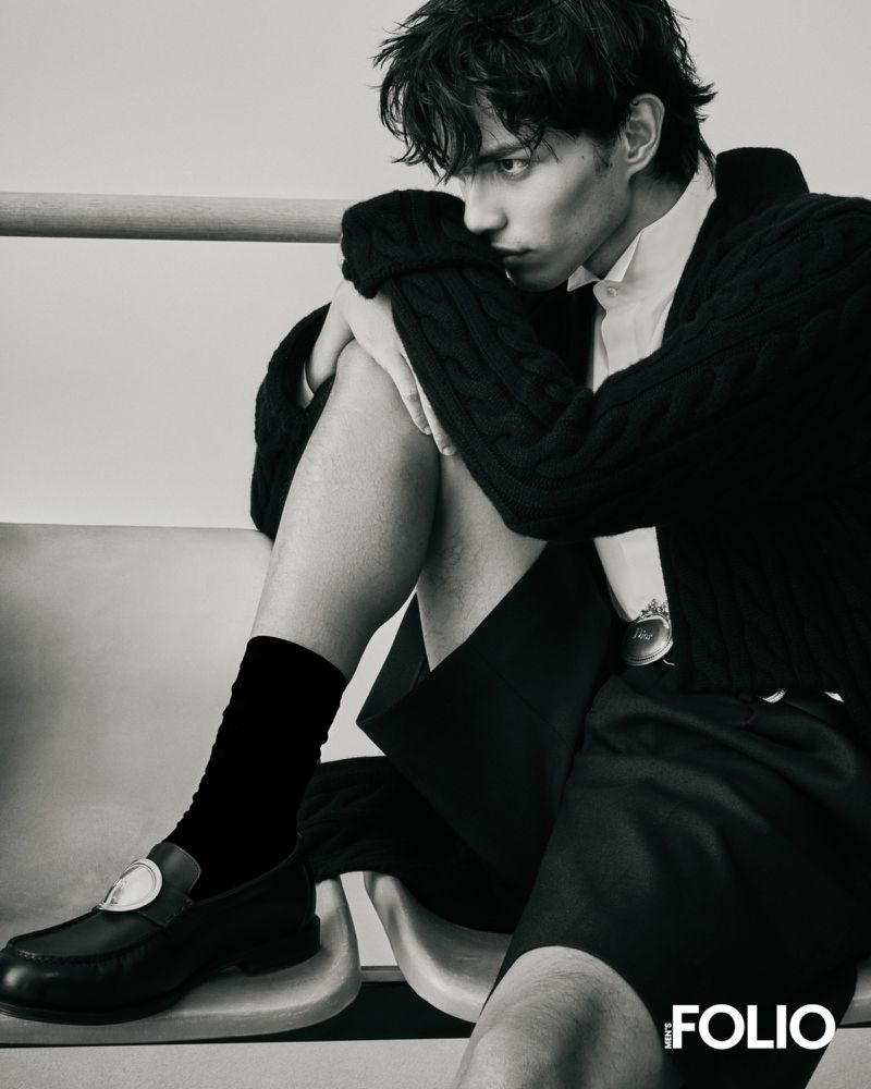 Model seated on a chair wearing a dark cardigan, shorts, and black loafers with white socks against a plain backdrop.