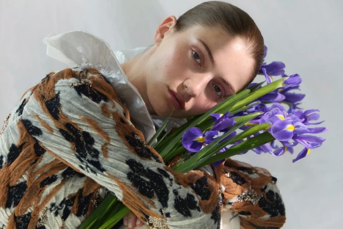 Model holding blue irises wearing a patterned brown top and white ruffled collar against a light background