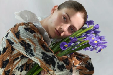 Model holding blue irises wearing a patterned brown top and white ruffled collar against a light background