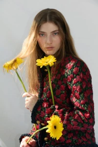Model with long blonde hair holding three yellow flowers wearing a red floral jacket against a white wall.