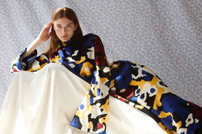 Model wearing a colourful patterned garment with a white draped skirt against a blue floral background