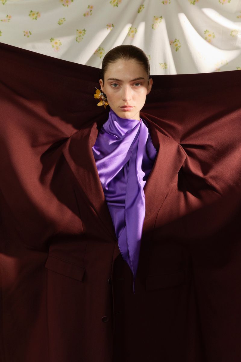 Model holding a burgundy garment outstretched, wearing a bright purple silk scarf and floral earring against a patterned backdrop.