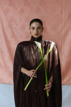 Model holding two calla lilies wearing a pleated brown garment against a pink patterned backdrop