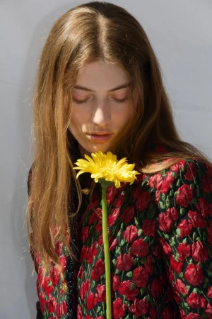 Close-up portrait of a woman with long brown hair holding a bright yellow gerbera daisy against a white wall.