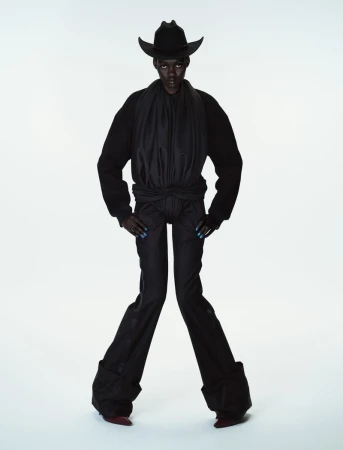 Model Caren Jepkemei in all-black attire including a cowboy hat and flared trousers against a plain white studio background.