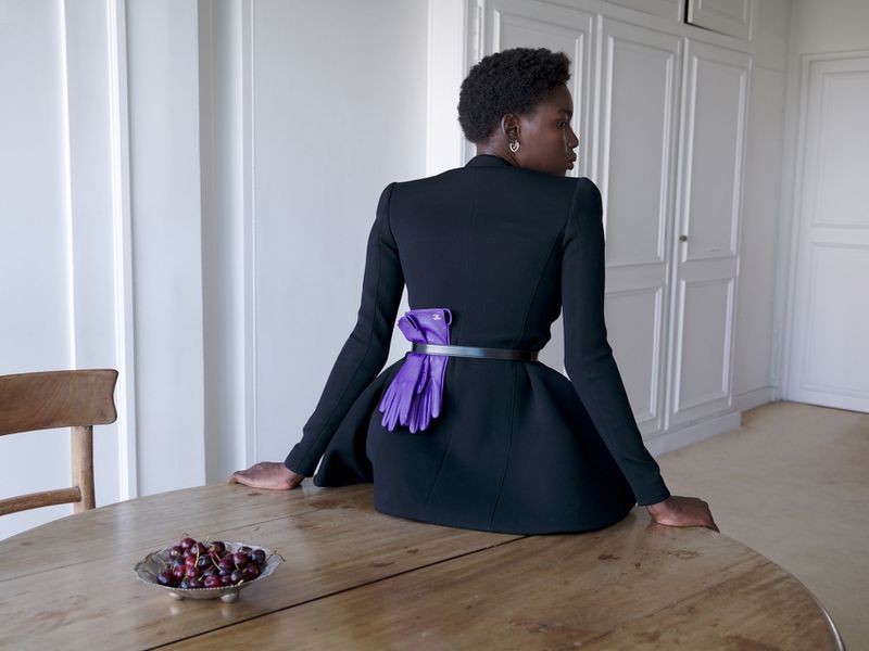 Model seated on a wooden table wearing a black jacket with purple gloves tucked into a belt for Annabelle Magazine Look Back.