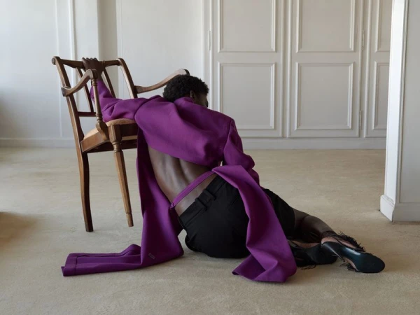 Model wearing a vibrant purple jacket and dark trousers leaning back against a wooden chair in a bright room.
