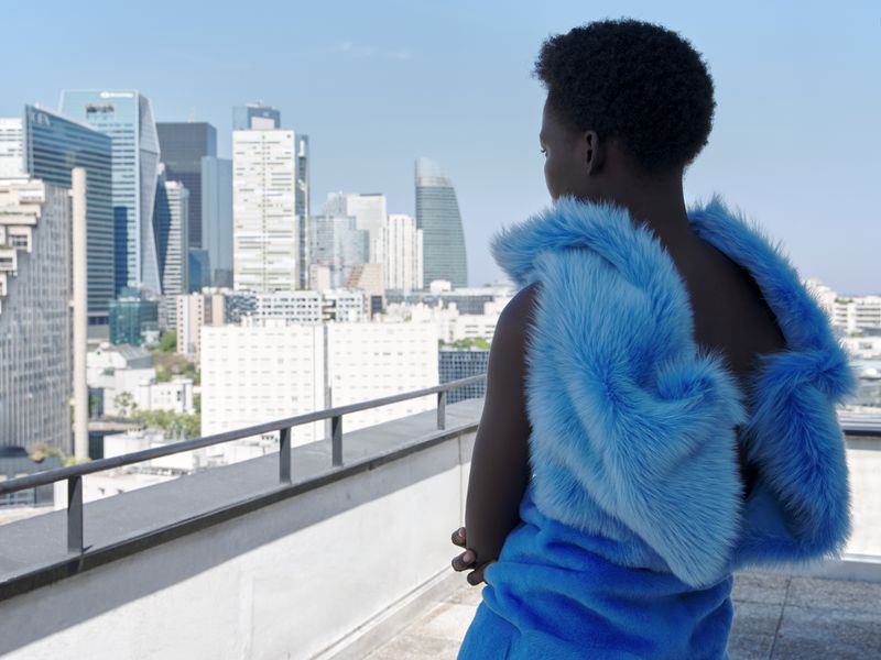 Model seen from behind wearing a bright blue faux fur garment on a rooftop overlooking a city skyline