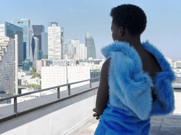 Model seen from behind wearing a bright blue faux fur garment on a rooftop overlooking a city skyline