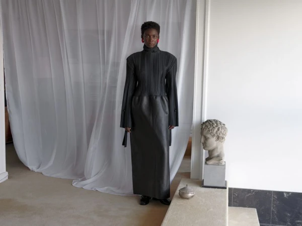 Model wearing a pleated black top and long skirt standing before white sheer curtains and a stone bust.