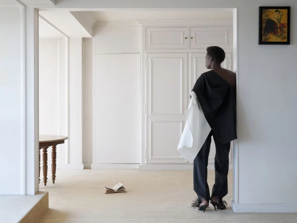 Model wearing asymmetric black and white draped top and dark trousers standing in a bright white apartment interior.