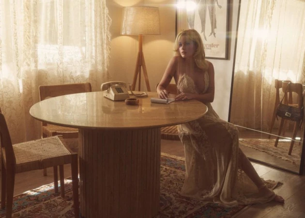 Woman in a long lace dress seated at a round wooden table in a retro living room with lace curtains and a floor lamp