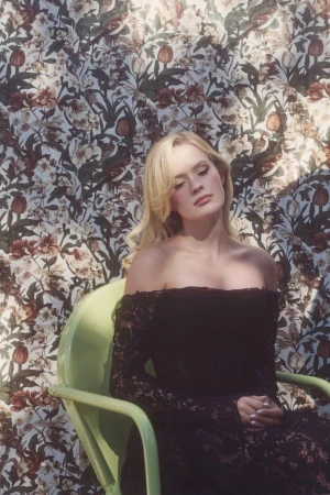 Blonde woman seated in a light green chair wearing a black off-the-shoulder lace dress against a floral wallpaper backdrop