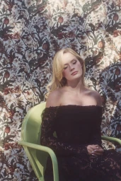 Blonde woman seated in a light green chair wearing a black off-the-shoulder lace dress against a floral wallpaper backdrop