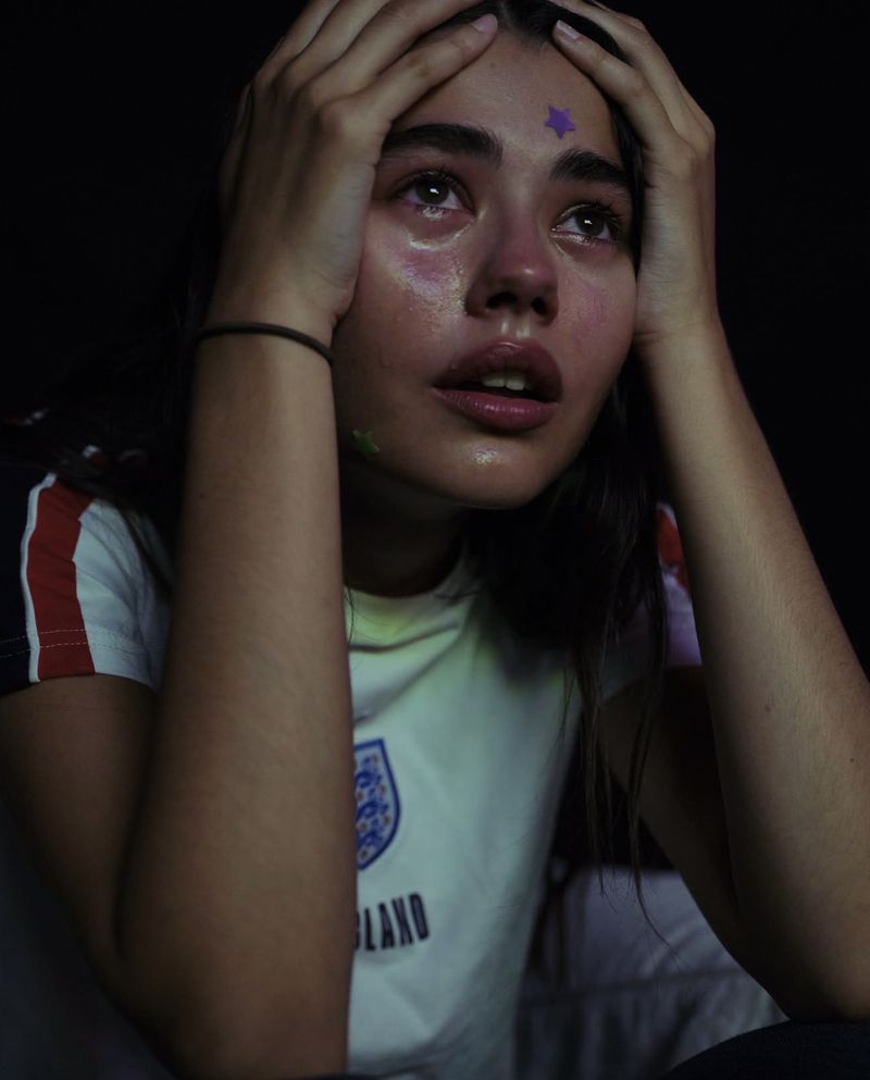 Close-up portrait of a young woman holding her head with hands, wearing a white t-shirt with red and blue stripes.