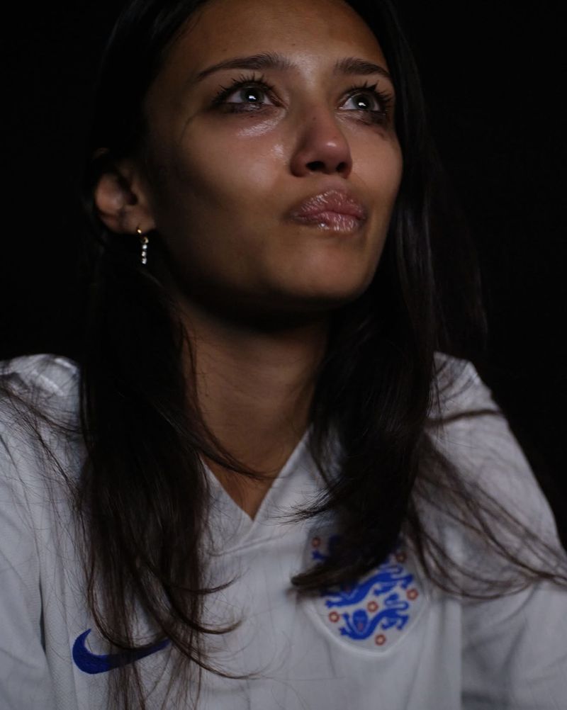 Close-up portrait of a young woman with dark hair wearing a white jersey looking upward against a dark background.