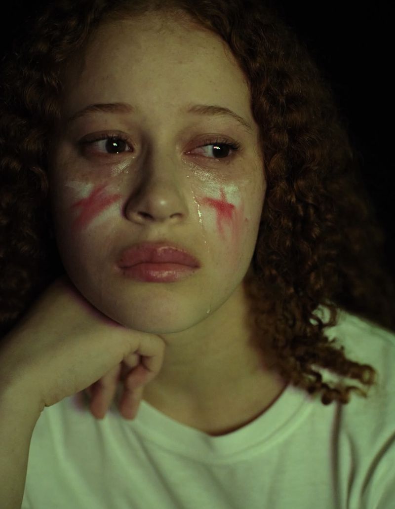 Close-up portrait of a young woman with curly brown hair, tear tracks, and painted red and white marks across her cheeks.