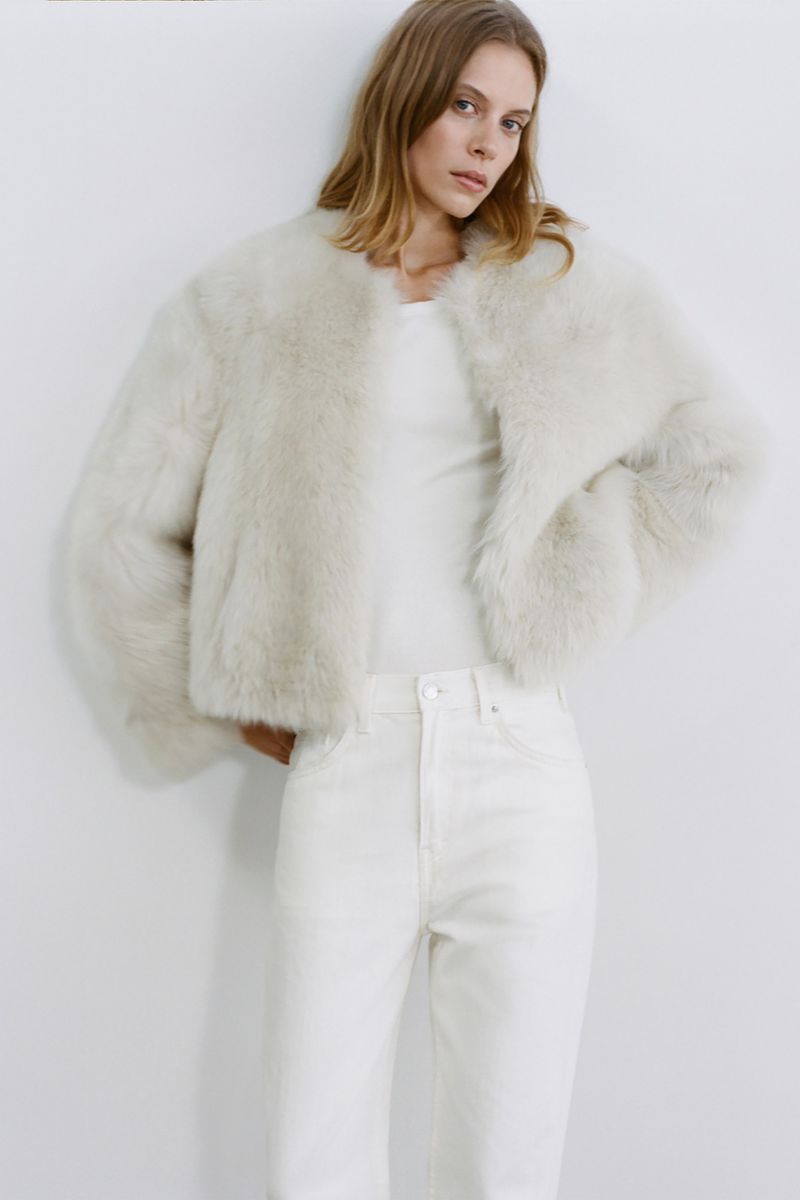 Fashion shot of a blonde woman in a white faux fur jacket and jeans, posing against a white backdrop in a bright, studio setting.