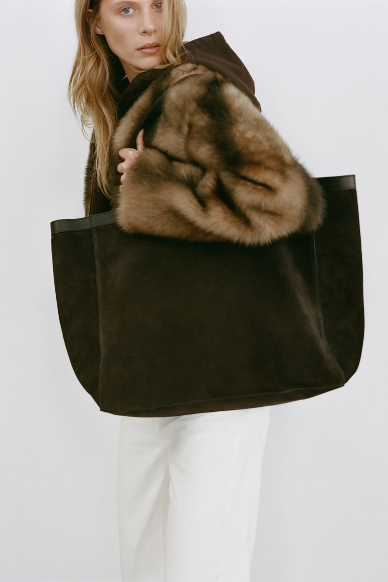 Woman poses with a brown tote bag and a fur-trimmed coat against a white background, showcasing fashion and style.