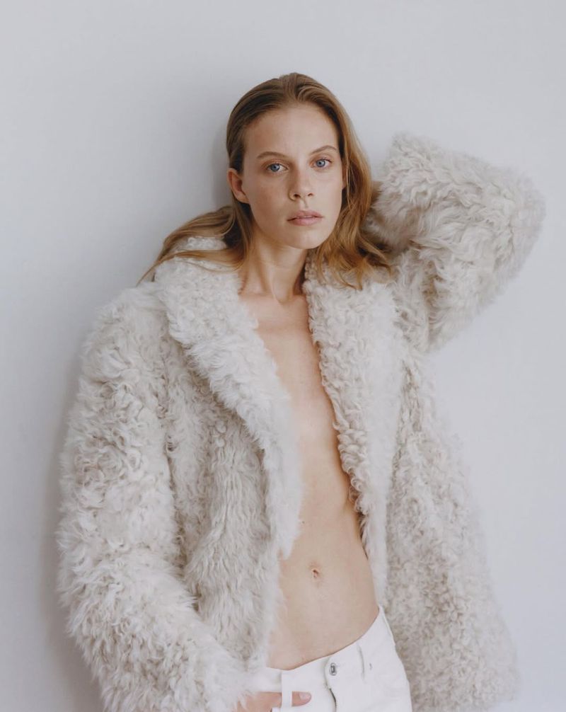 A woman with blonde hair poses in an unbuttoned, light-beige, furry coat and white pants against a neutral wall, looking at the camera.