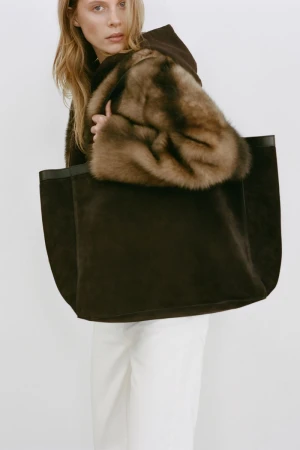 Model Anouk Smits carrying an oversized dark brown suede tote bag with fur trim for Nili Lotan Resort 2026 against a white backdrop.