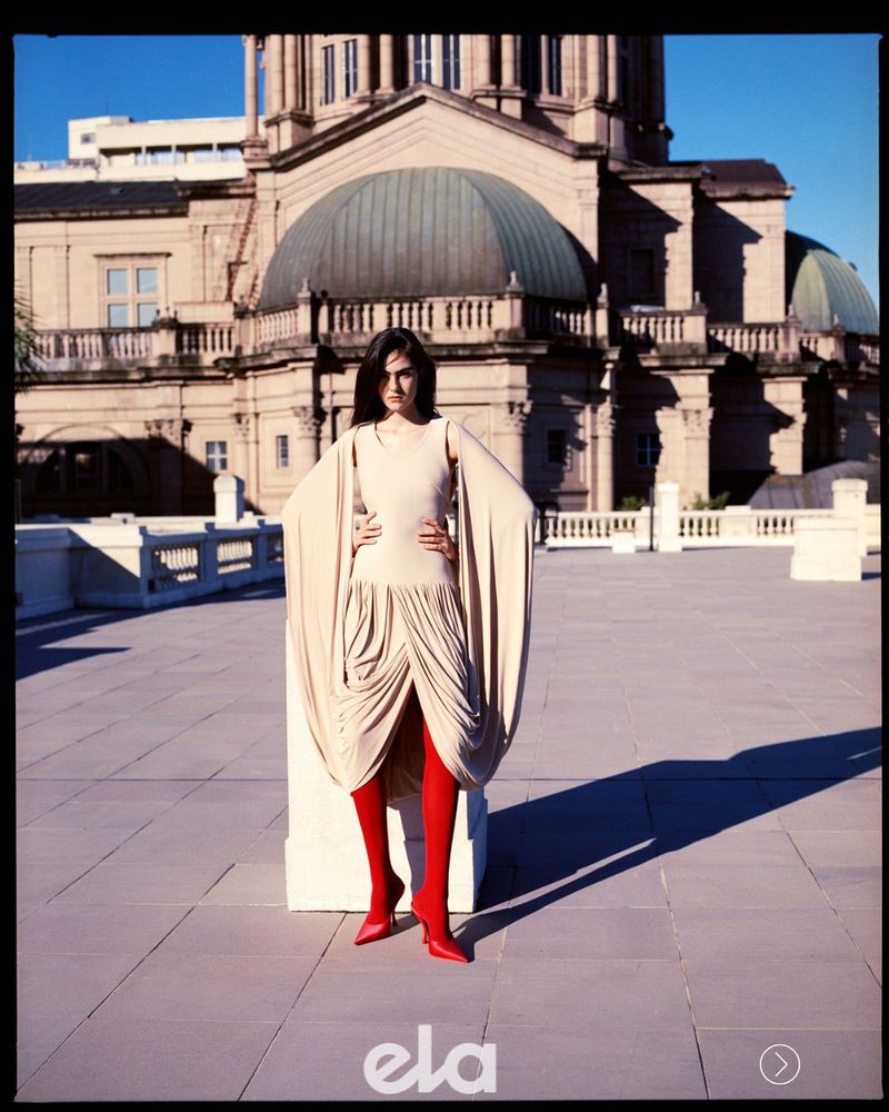 Model wearing a draped beige dress and bright red tights standing before a classical domed building