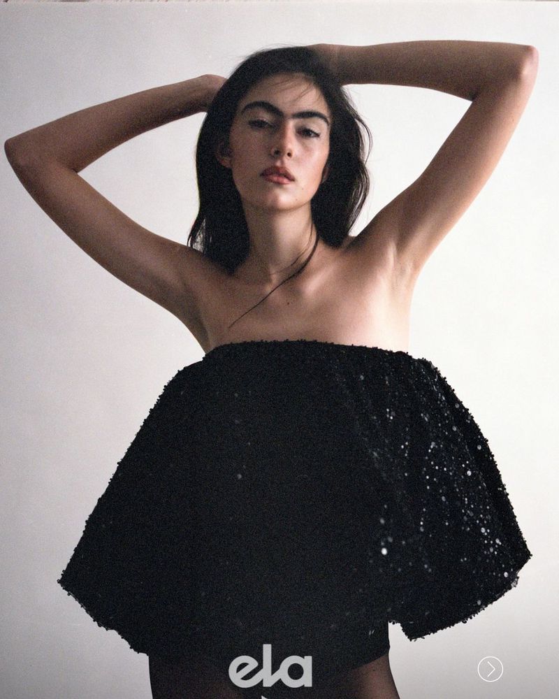 Woman wearing a strapless black sequin dress with hands behind her head against a plain light background