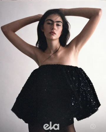 Woman wearing a strapless black sequin dress with hands behind her head against a plain light background