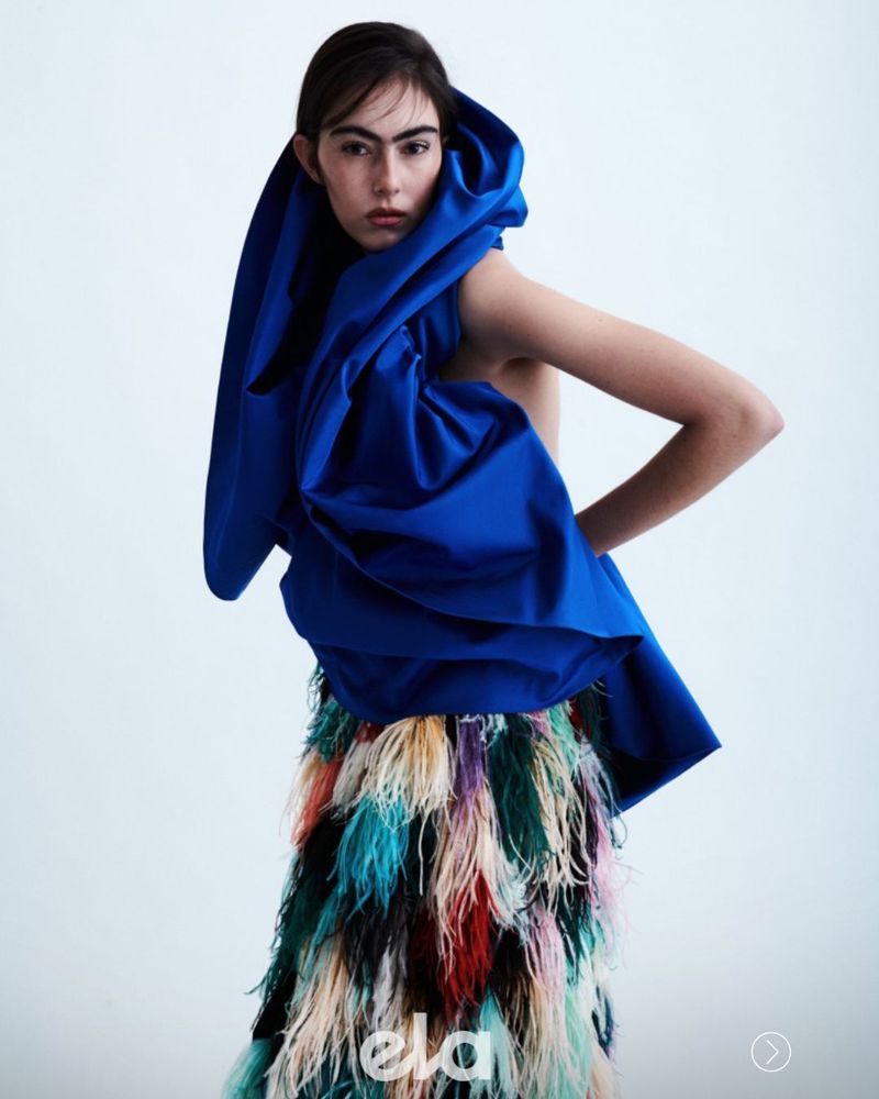Model wearing a structured royal blue hooded top and a colorful feathered skirt against a white studio background.
