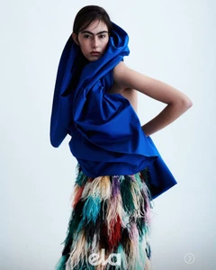 Model wearing a structured royal blue hooded top and a colorful feathered skirt against a white studio background.